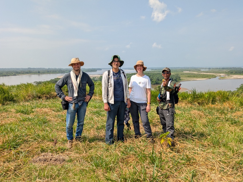 Four people stand together in a grassy lakeside landscape on a sunny day
